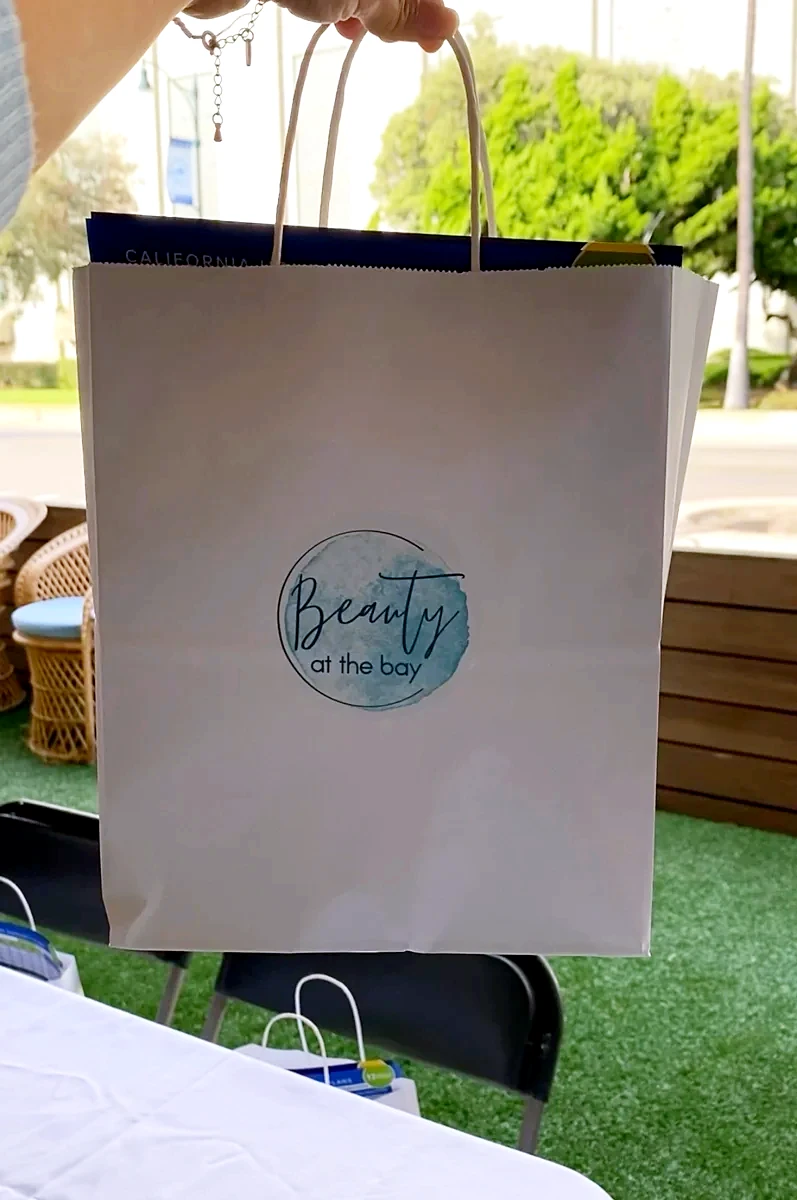 White Beauty at the Bay goodie bag with the watercolor logo, held up at an outdoor event
