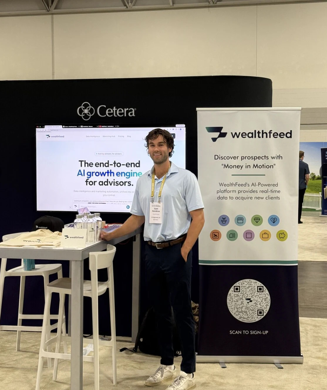 WealthFeed booth at an indoor conference with a retractable banner reading Discover prospects with Money in Motion, event icons and a scan-to-sign-up QR code