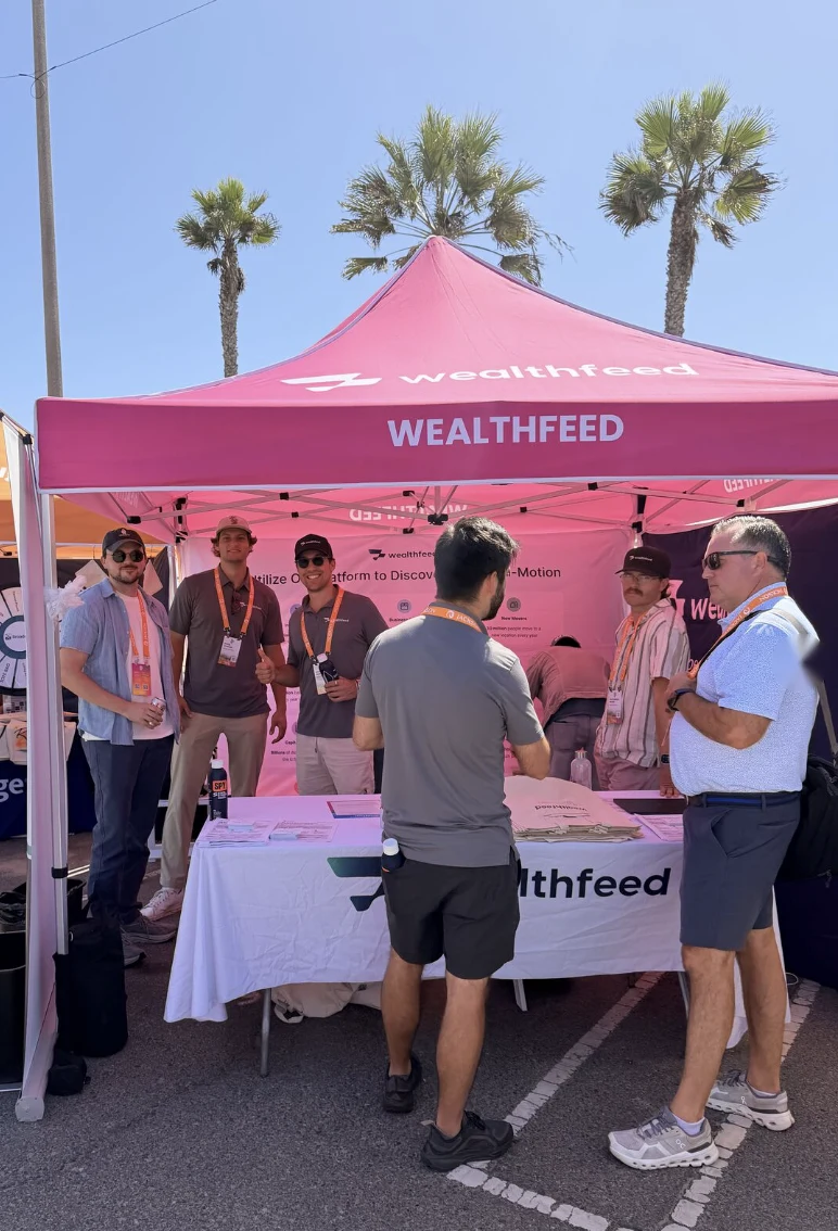 The pink WealthFeed tent at an outdoor conference under palm trees, with the team at a branded table and the side wall behind them